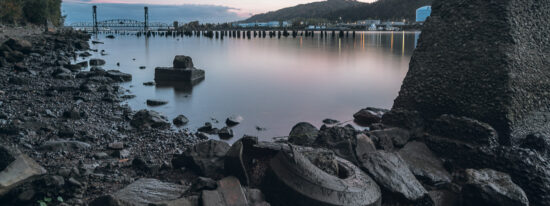 The rocky bank of the Willamette River near St. John's. The train bridge is in the distance and an old tire blends in with the rocks Superfund cleanup